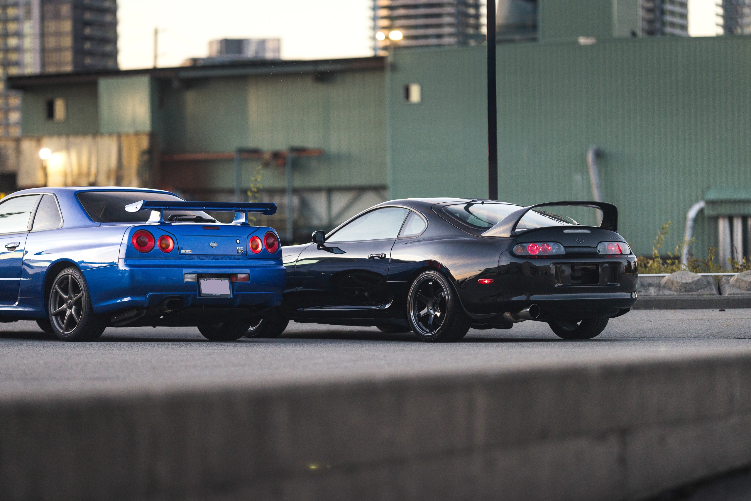 The black Supra rear three-quarter view beside the R34, framed over a concrete wall